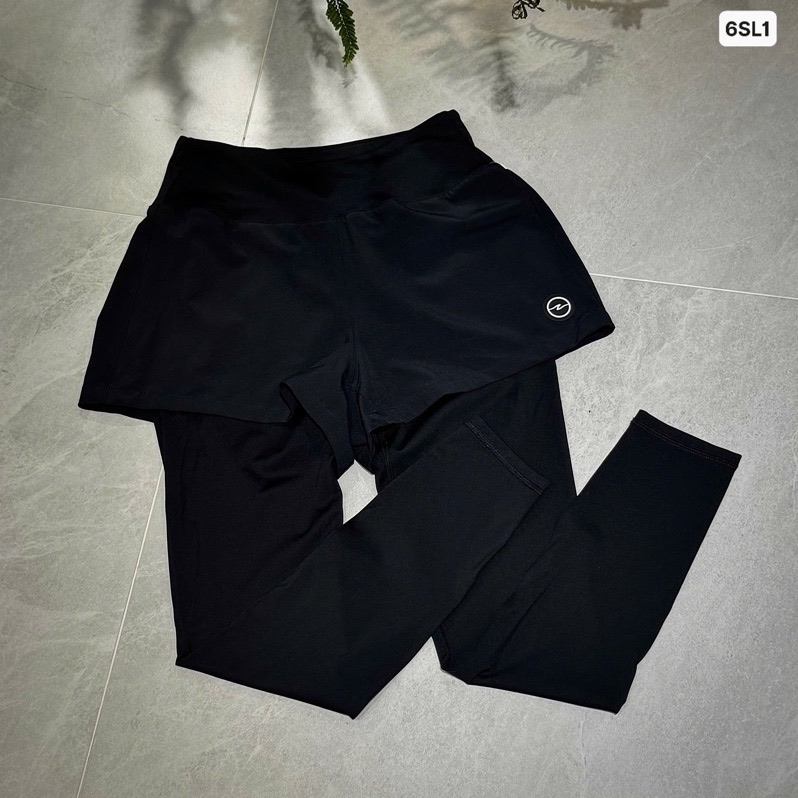 Quần legging đen liền quần short bên ngoài, cạp to bản, logo tròn nhỏ ở ống short, trải phẳng trên nền đá xám