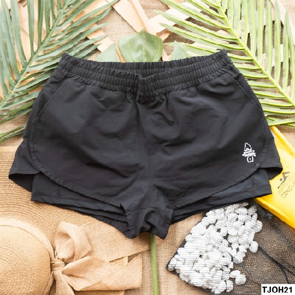 Quần short nữ màu đen hai lớp, cạp chun, logo in trắng ở ống, trải phẳng trên nền cói với lá cọ và mũ cói